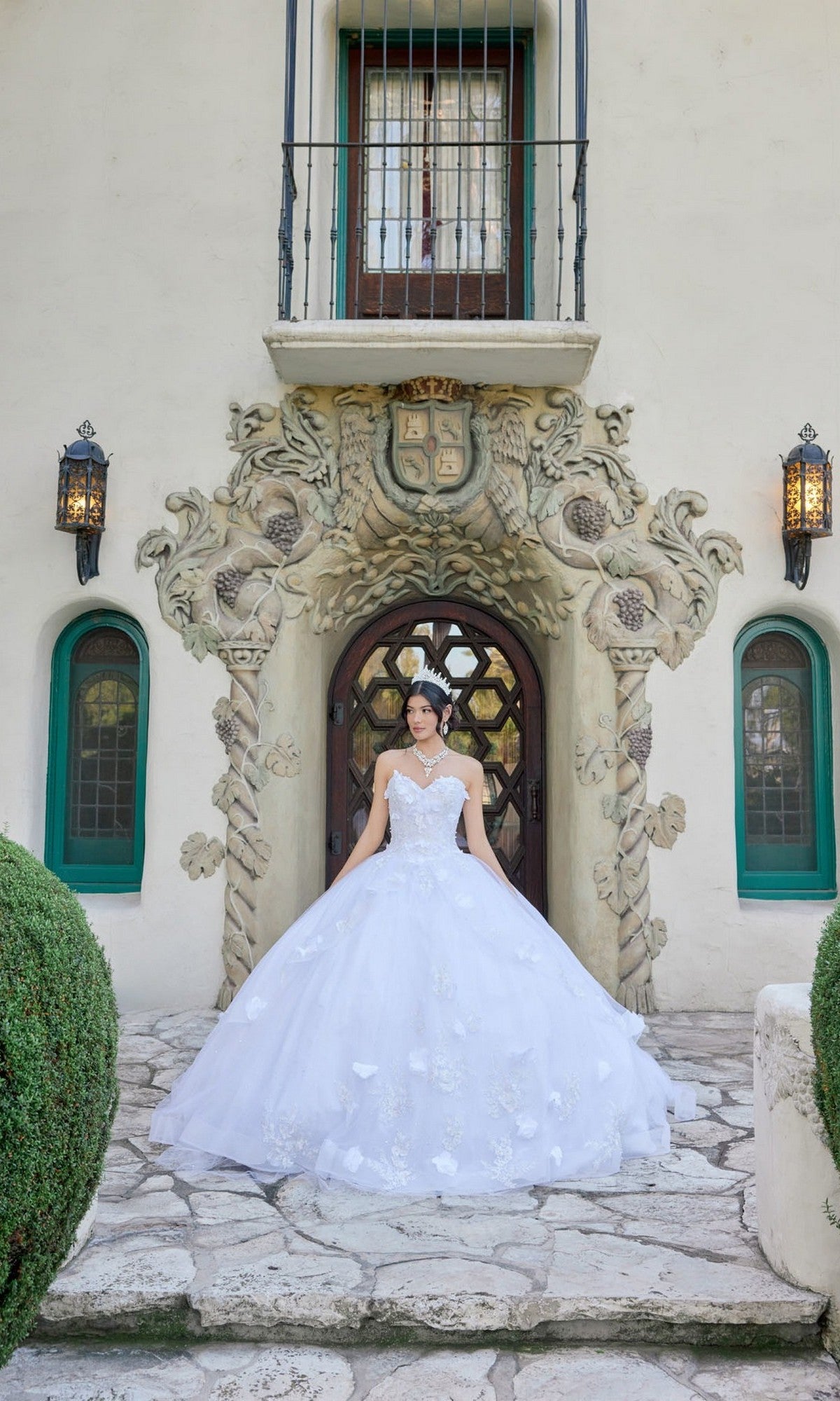 Wearing the Long Wedding Dress: Juliet JT1495HW—an elegant strapless white ball gown with a sweetheart neckline and delicate floral appliques—a woman stands before an ornate doorway featuring intricate stonework and green-trimmed windows.